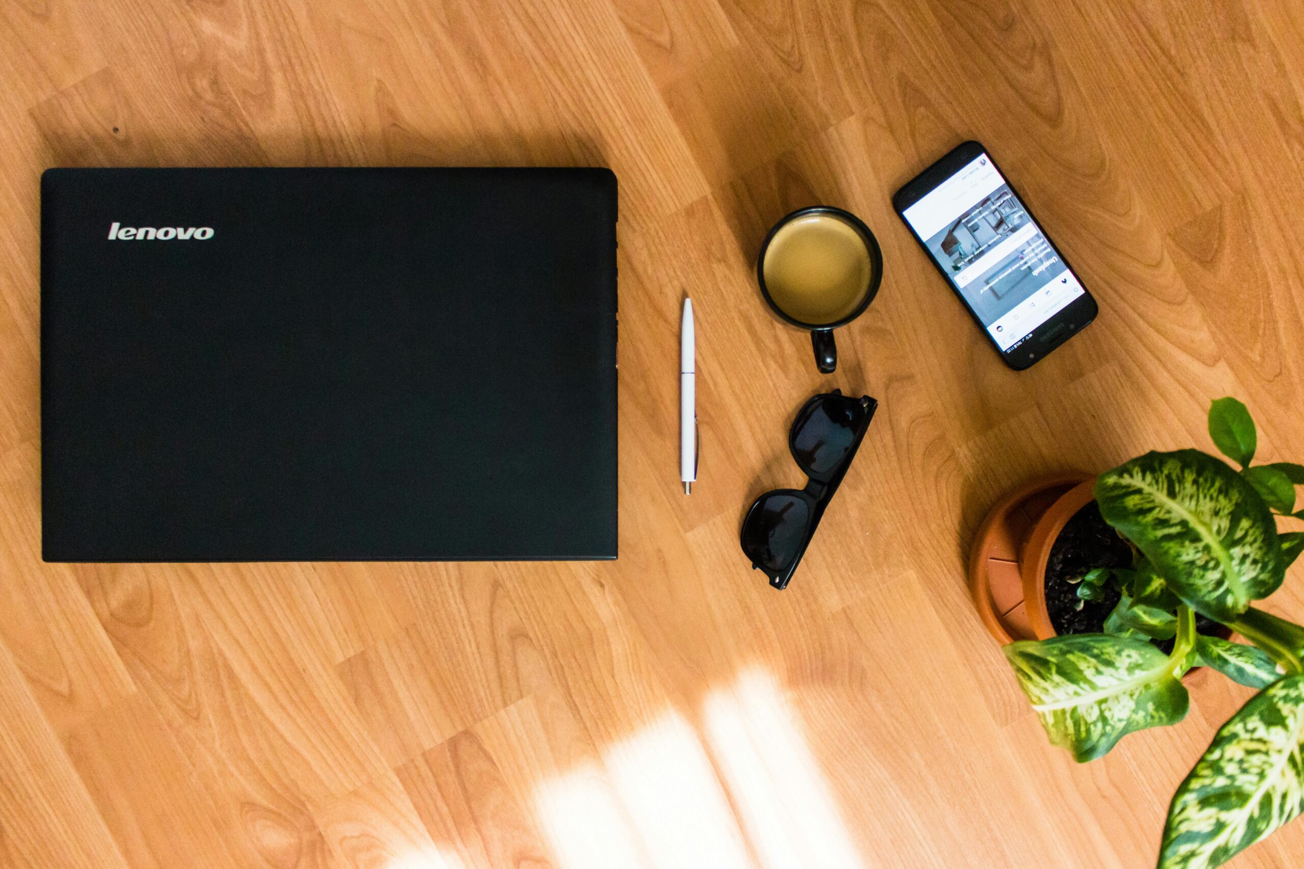 Lenovo laptop on wood tabletop beside pen, tablet, coffee, sunglasses, and a potted plant 